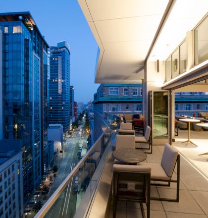 A modern cityscape with tall buildings and a stylish balcony featuring tables and chairs, overlooking a street at dusk.