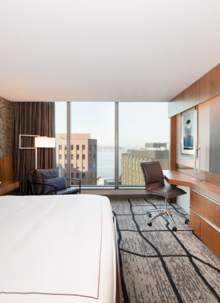 A modern hotel room with a large bed, wall-mounted TV, desk, and a window view of city buildings and water.