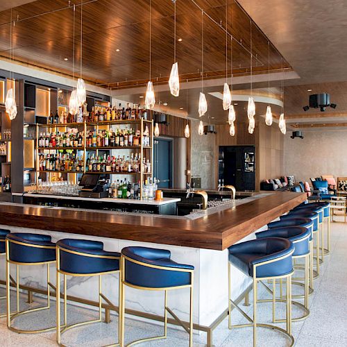 A modern bar with blue chairs, hanging lights, and a stocked liquor shelf in a spacious lounge area with seating and tables.