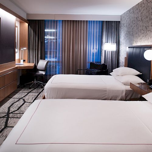 A modern hotel room with two beds, a TV, desk, and chair; patterned wallpaper and carpet add style, while lamps provide warm lighting.