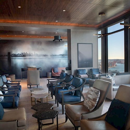 A modern lounge with plush seating, large windows with a waterfront view, and a skyline mural on the wall, creating a relaxed atmosphere.