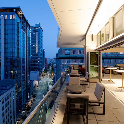 A modern rooftop terrace with seating overlooks a city's high-rise buildings and streets at dusk, featuring stylish architecture.