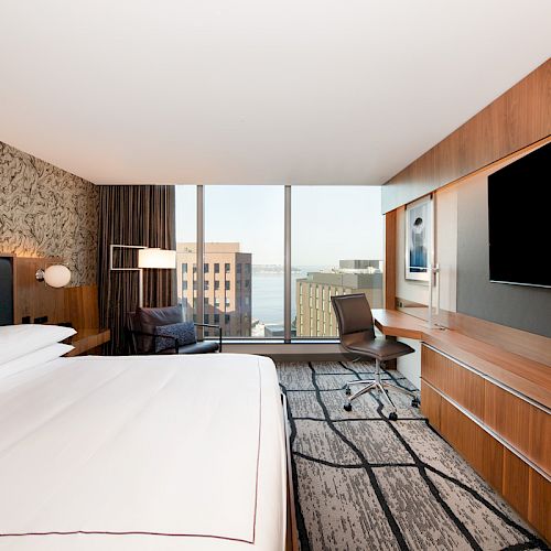 A modern hotel room with a large bed, wall-mounted TV, desk, chair, and a window view of buildings.