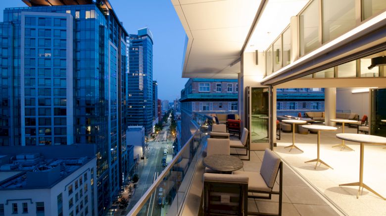 A modern city skyline at dusk with high-rise buildings, and a balcony featuring tables and chairs overlooking the street below.