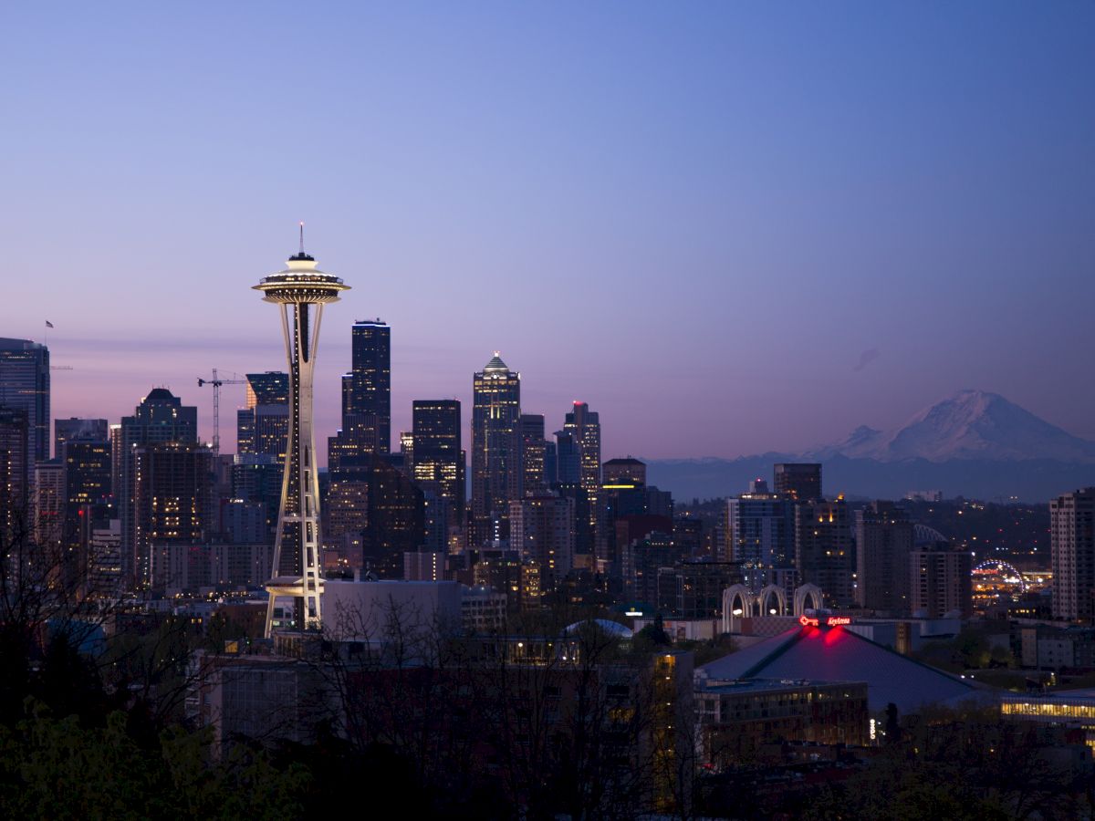 A cityscape at dusk with a prominent tower, part of downtown, and a distant mountain silhouette against a blue and purple sky.