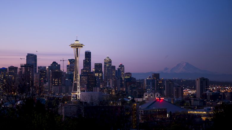 A cityscape at dusk with a prominent tower, part of downtown, and a distant mountain silhouette against a blue and purple sky.