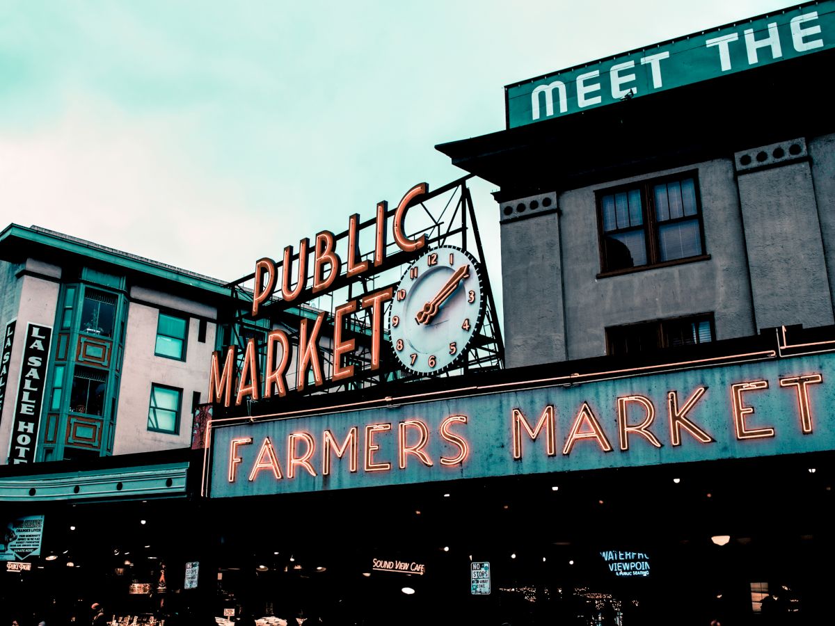The image shows a neon sign for a public farmers market with a clock, set against buildings with an overcast sky in the background.