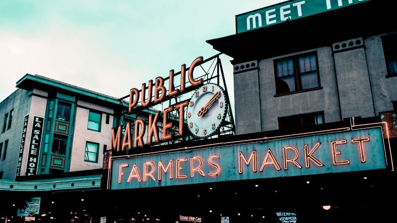 The image shows a neon sign for a public farmers market with a clock, set against buildings with an overcast sky in the background.