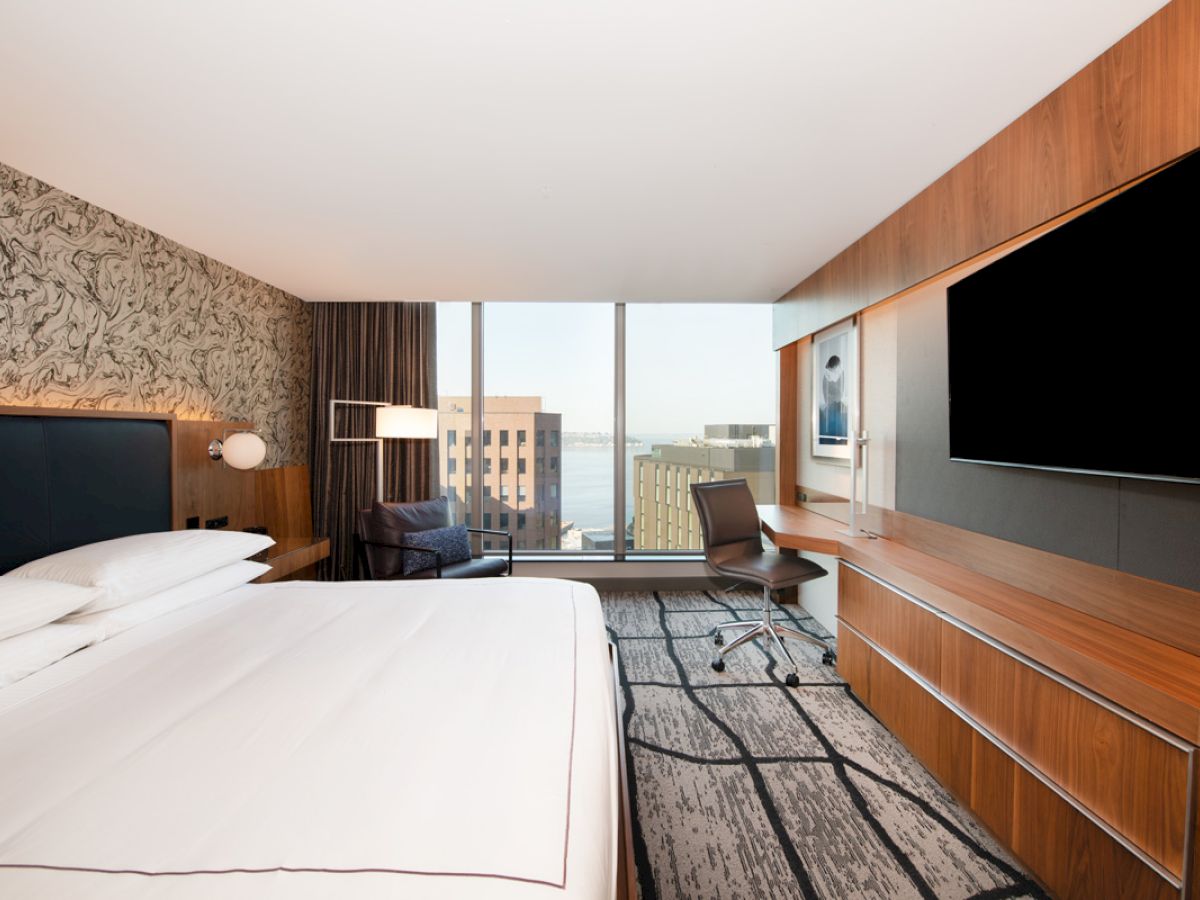 A modern hotel room features a bed, large TV, desk, and city view through expansive windows, blending comfort with urban style.