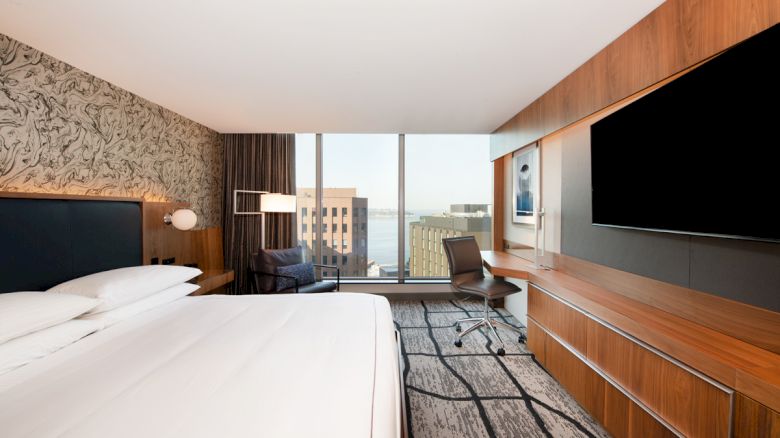 A modern hotel room features a bed, large TV, desk, and city view through expansive windows, blending comfort with urban style.
