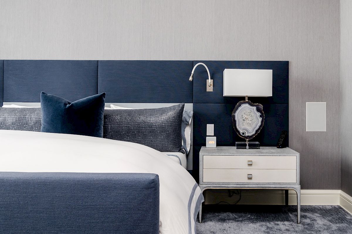 A modern bedroom features a navy and white color scheme with a bed, nightstand, lamp, and plush carpet, creating a sleek, cozy atmosphere.