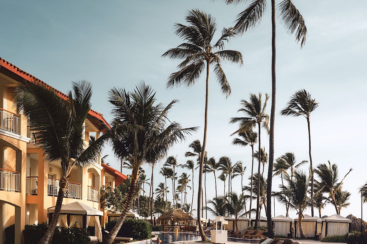 A resort pool area with palm trees, lounge chairs, and a building in the background under a clear sky.