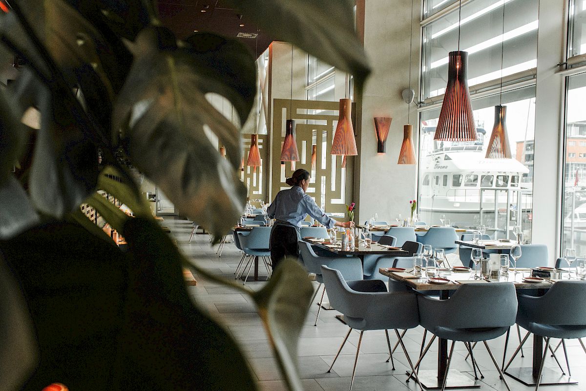 A restaurant with modern decor, featuring a worker setting tables. Large plants and pendant lights give a stylish ambiance.