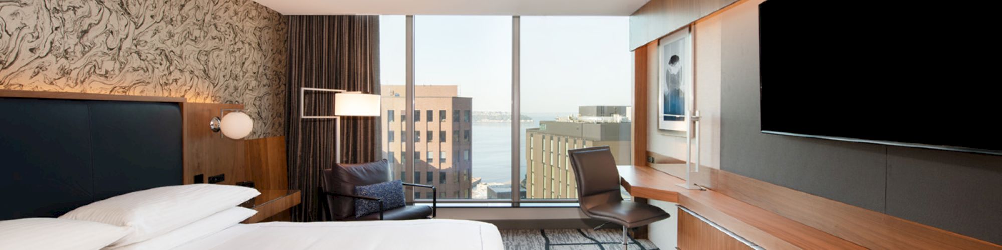 A modern hotel room with a large bed, desk, chair, wall-mounted TV, and a city view through floor-to-ceiling windows completes the scene.